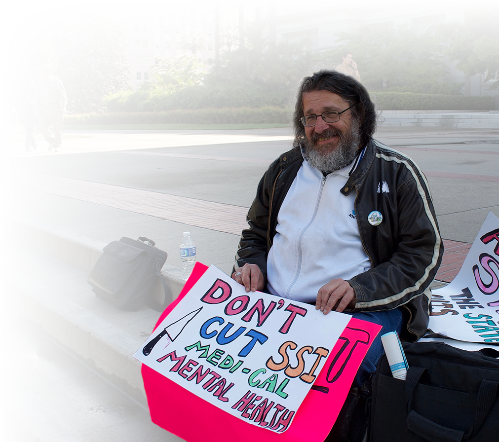 Photo of a man on the steps of the Capitol with a sign saying, "Don't cut SSI, Medi-Cal, and Mental Health (Benefits)."